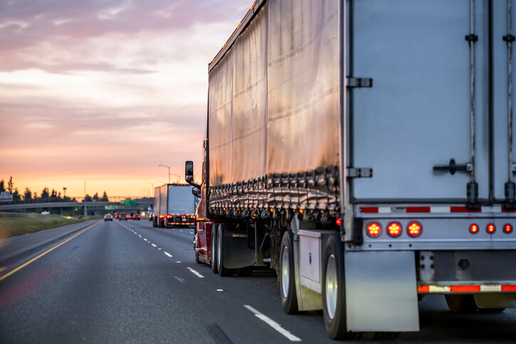 Burgundy big rig semi truck with black covered semi trailer driving on the highway road at twilight with pink clouds sky
